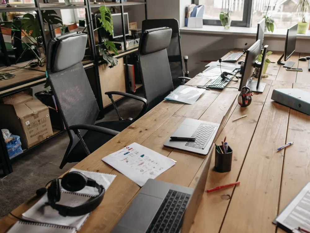 Shared office workspace with laptops, monitors and headsets on a long wooden desk